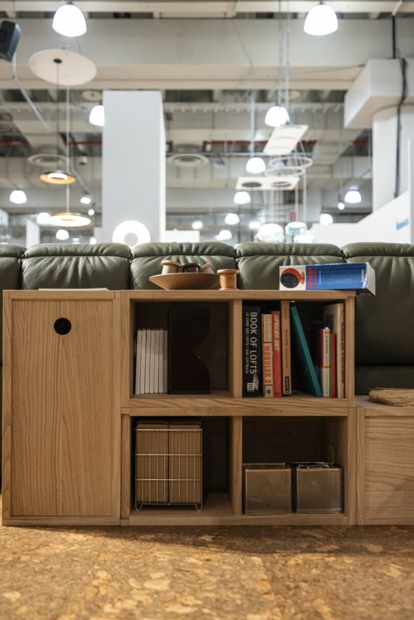 A wooden shelving unit with books, storage boxes, and decorative items is placed in front of a green sofa in a well-lit room with ceiling lights
