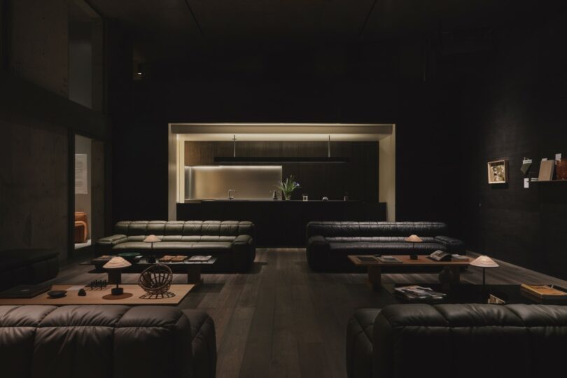 A dimly lit modern living room with dark sofas, low wooden tables, decorative lamps, and an open kitchen in the background