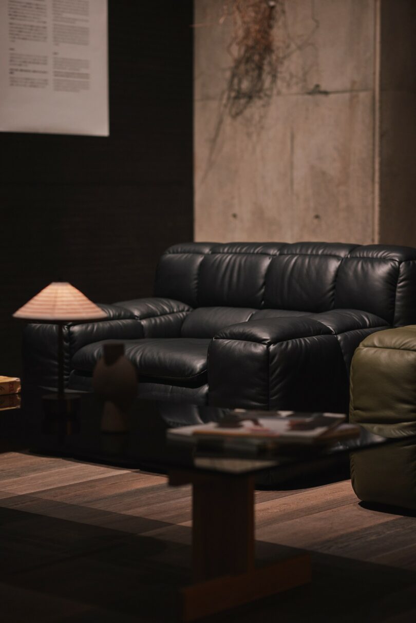 A black leather sofa sits beside a low table with magazines and a small lamp in a dimly lit modern living room