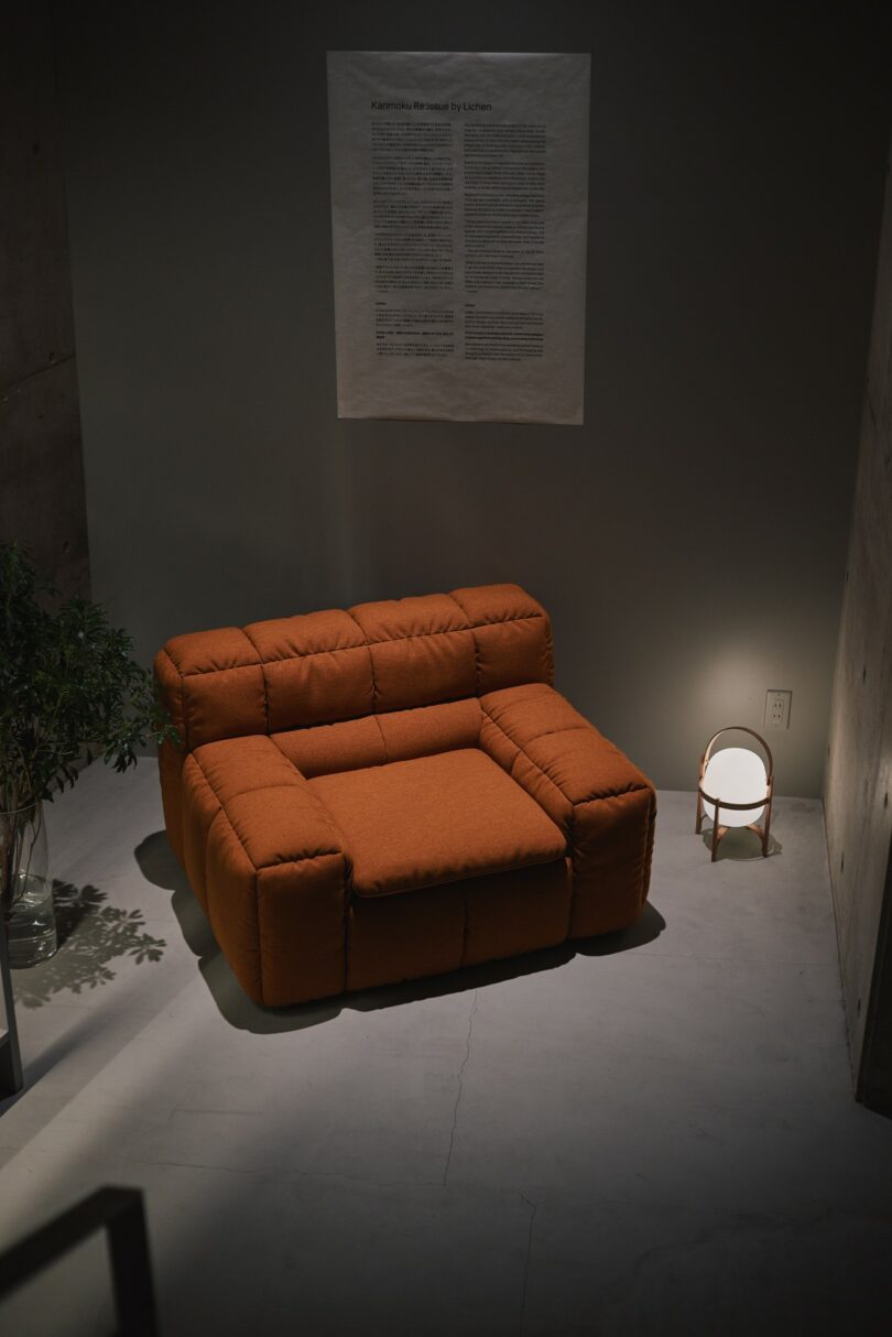 An orange upholstered armchair sits on a light floor next to a small round lamp and a potted plant; a text poster hangs on the wall behind it