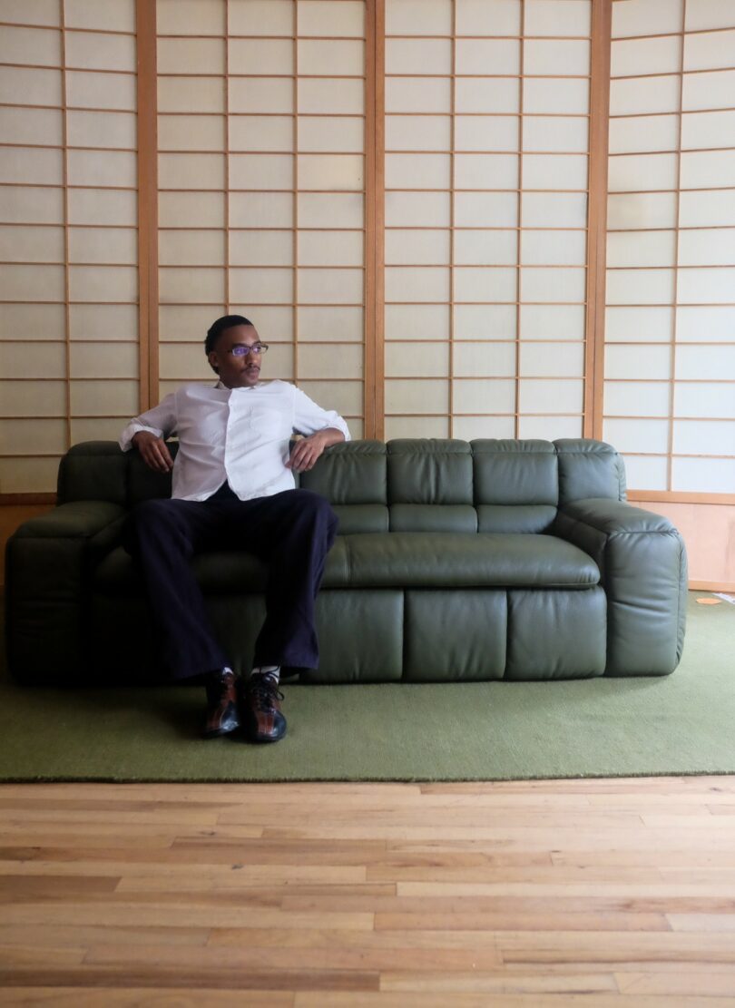 A person in a white shirt and dark pants sits on a green sofa in a room with wooden floors and shoji screen walls