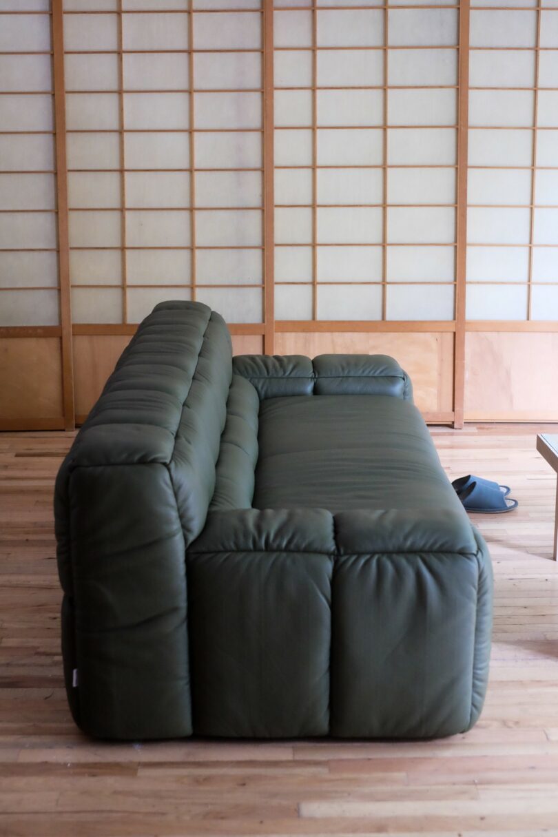 A dark green, tufted sofa is placed on a wooden floor in front of shoji screen panels in a minimalist room