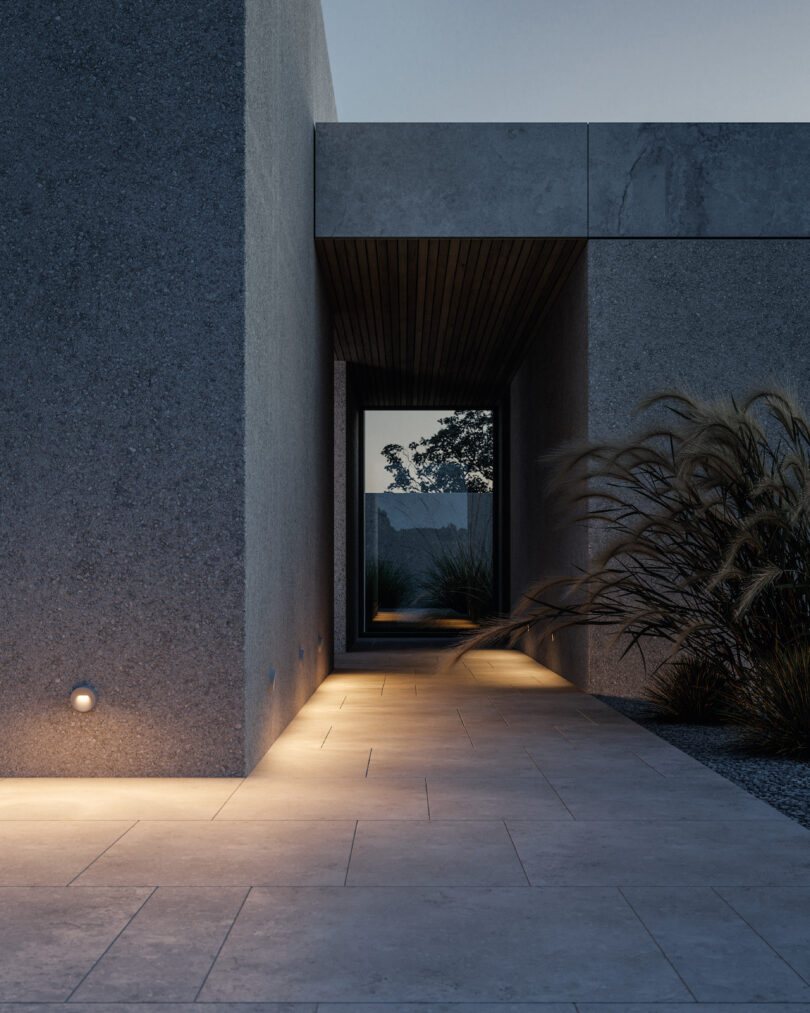 A modern concrete building with a tiled walkway, recessed lighting, and tall grasses, leading to an open corridor framed by a large rectangular doorway