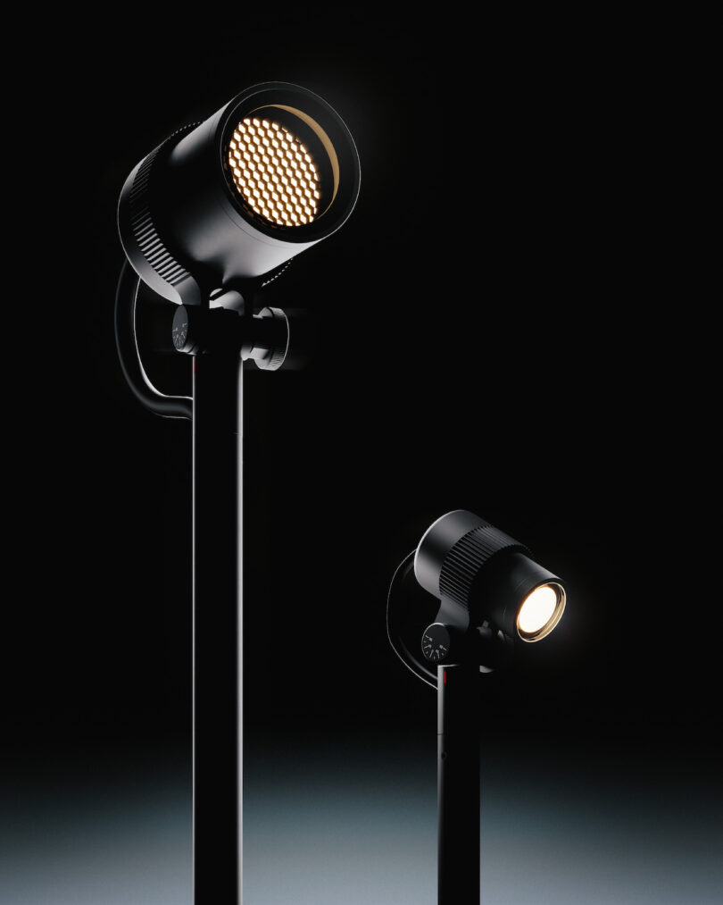 Two black stage spotlights on stands are illuminated against a dark background, casting soft light