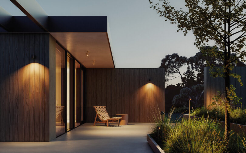 A modern patio at dusk featuring wooden walls, outdoor lighting, a woven lounge chair, a small table, and landscaped plants next to a tree