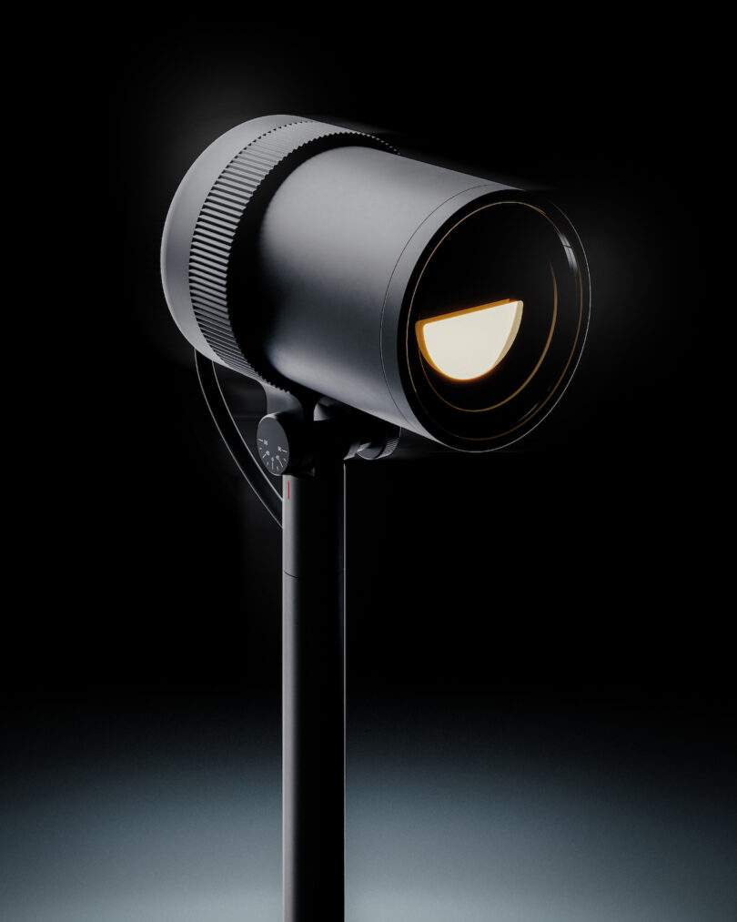 A modern black spotlight with a cylindrical design is mounted on a vertical stand, illuminated against a dark background