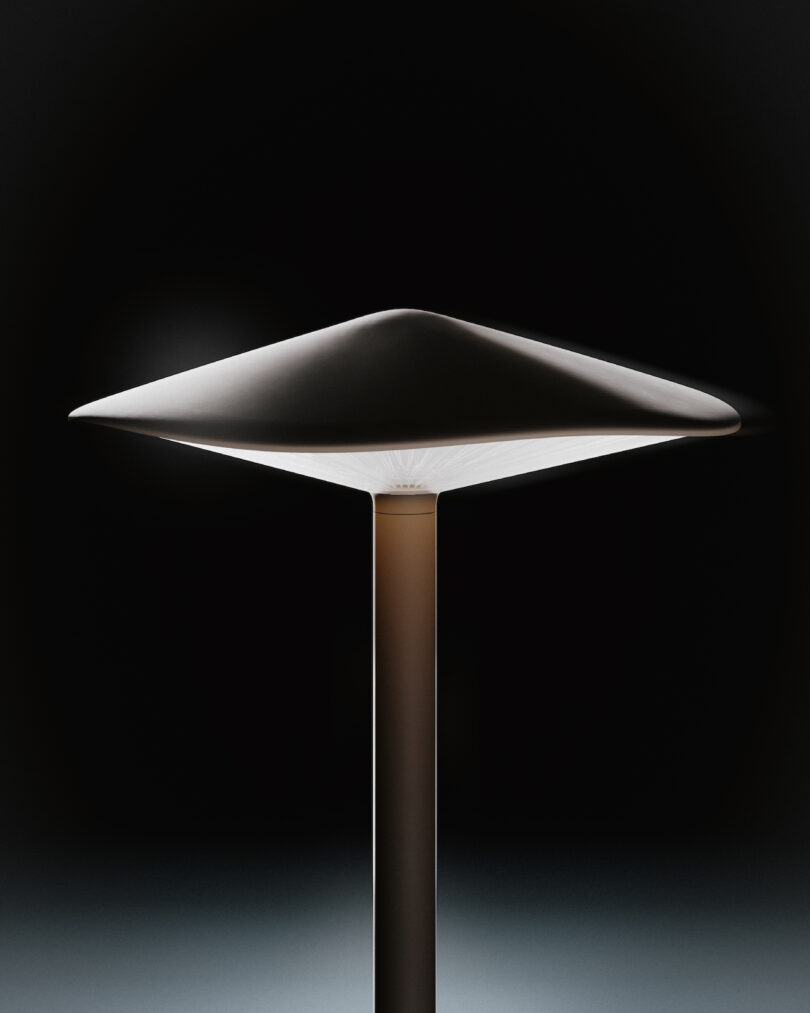 A modern, minimalist outdoor light with a wide, flat, triangular shade on a central cylindrical stand, illuminated against a dark background
