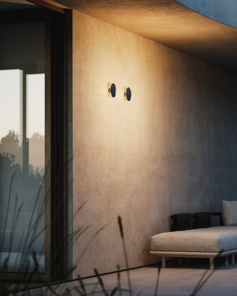 A modern outdoor patio with a beige lounge chair against a smooth stucco wall, illuminated by two round wall lights at dusk