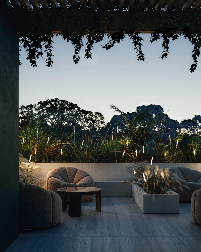 A modern outdoor lounge at dusk with cushioned chairs, a small table, decorative grasses, soft lighting, and overhead vines. Trees are visible in the background