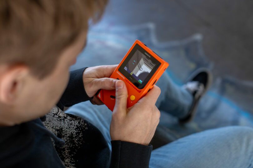A person holding and playing a small orange handheld gaming device, viewed from above.