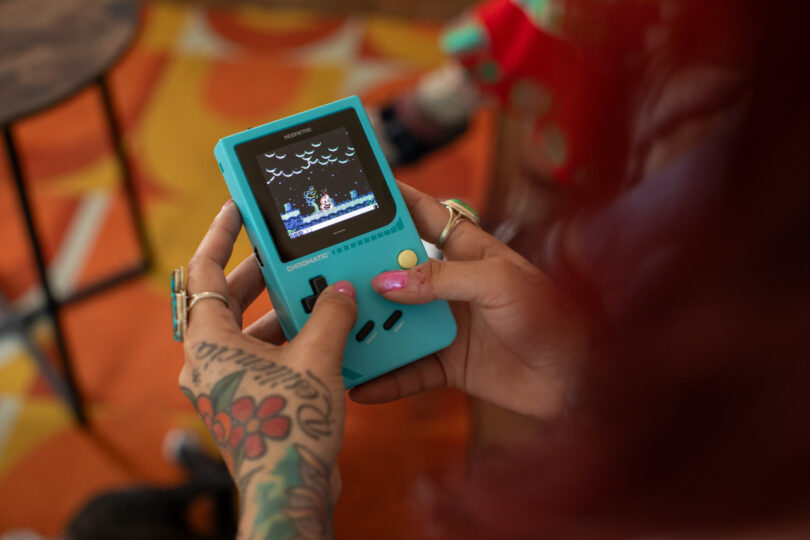 A person with tattoos and rings holds a turquoise handheld gaming console, playing a retro video game.