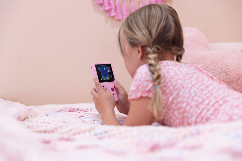 A young girl with braided hair lies on a bed, holding and looking at a pink handheld gaming device.