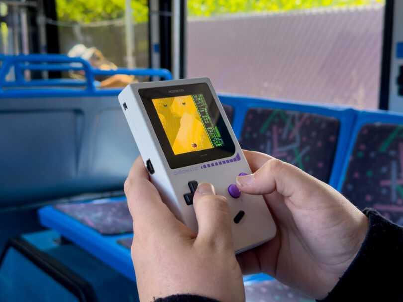 Person holding and playing a handheld video game console with a desert-themed game displayed, seated on a blue bench seat inside a bus.