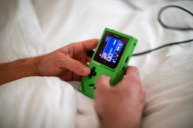 Person holding a green handheld gaming console and playing a block-stacking game while sitting on a white blanket.