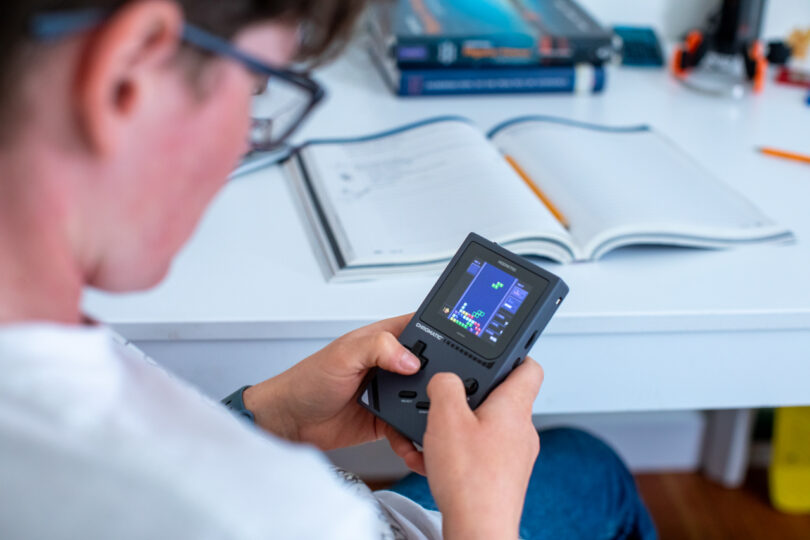 A person holds and plays a handheld video game console showing a puzzle game, with an open notebook and pencil on the desk in the background.