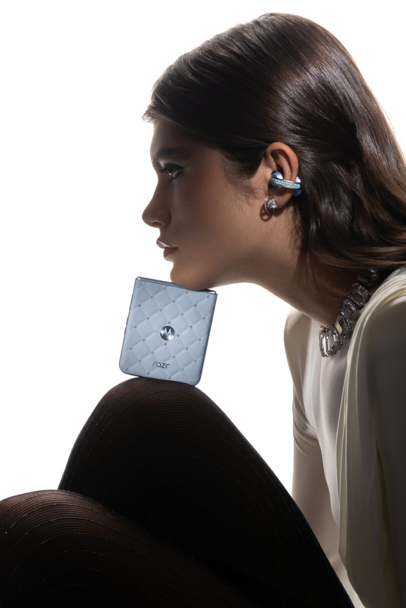 Woman in profile wearing a white top, black tights, chunky necklace, and wireless earbud, balancing a silver Motorola Razr phone on her chin against a white background.