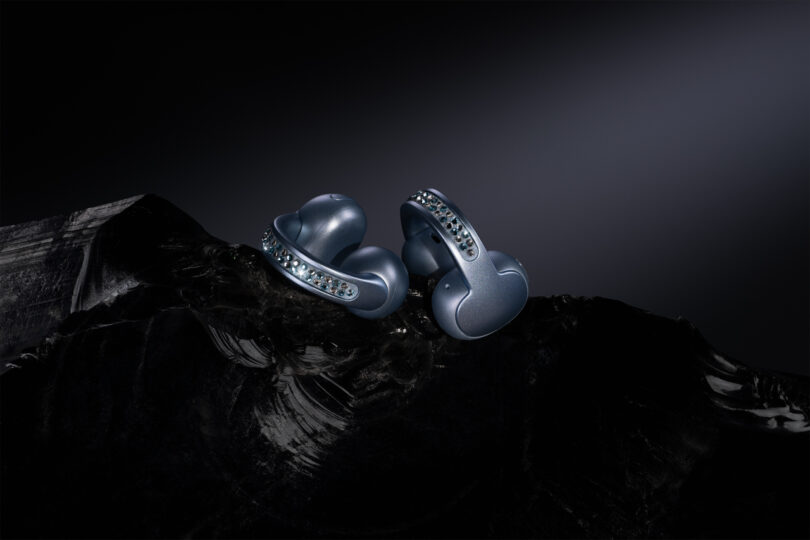 A pair of metallic blue wireless earbuds with sleek, curved design and button details displayed on a dark, textured surface against a black background.