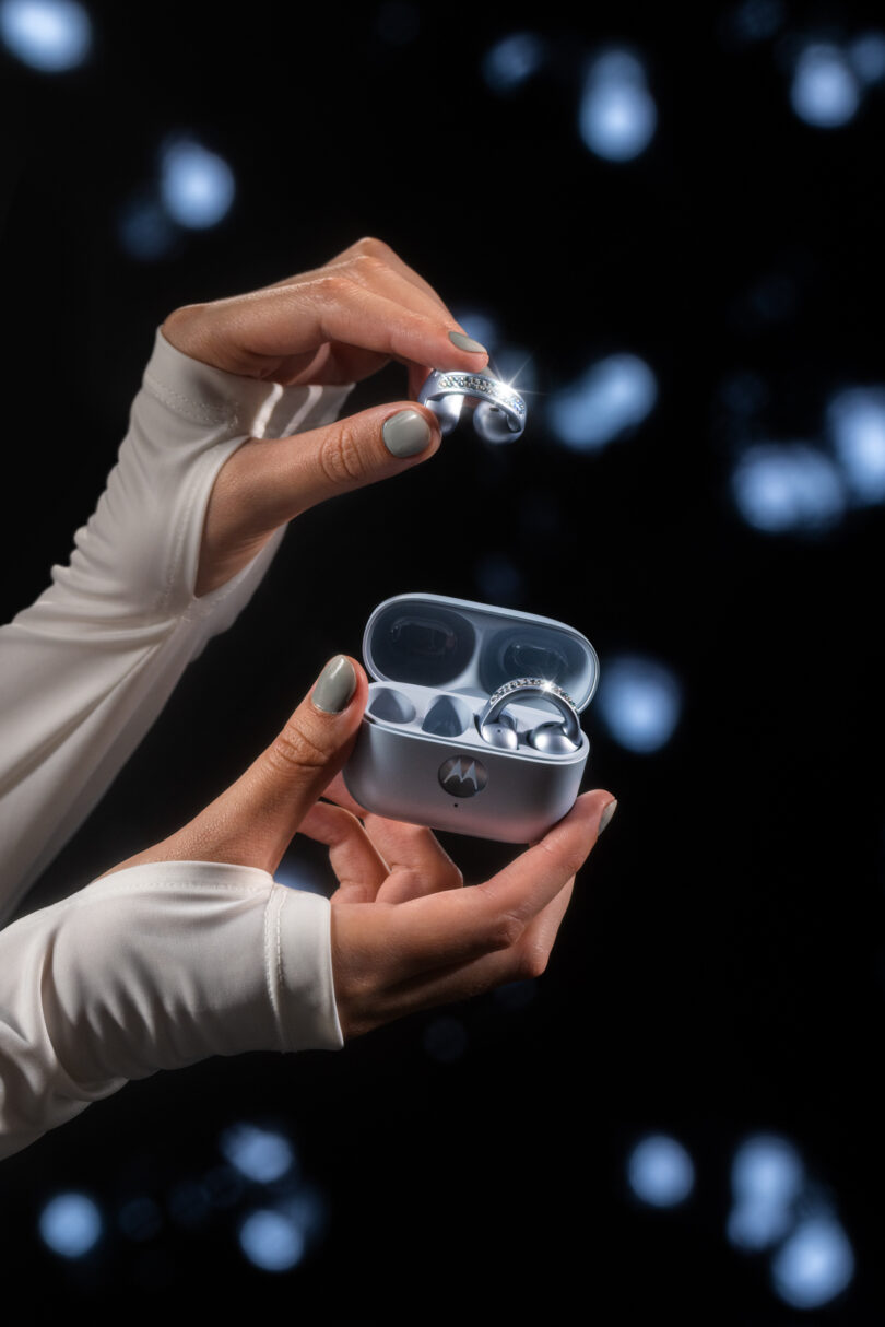 A person wearing white sleeves holds a Motorola wireless earbud and charging case against a dark background with bokeh lights.