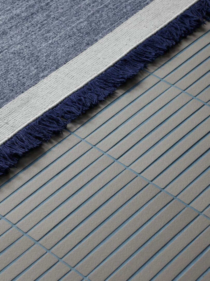 Primer plano de una alfombra con flecos azules y blancos que cubre parcialmente el suelo de baldosas grises con finas líneas de lechada azul