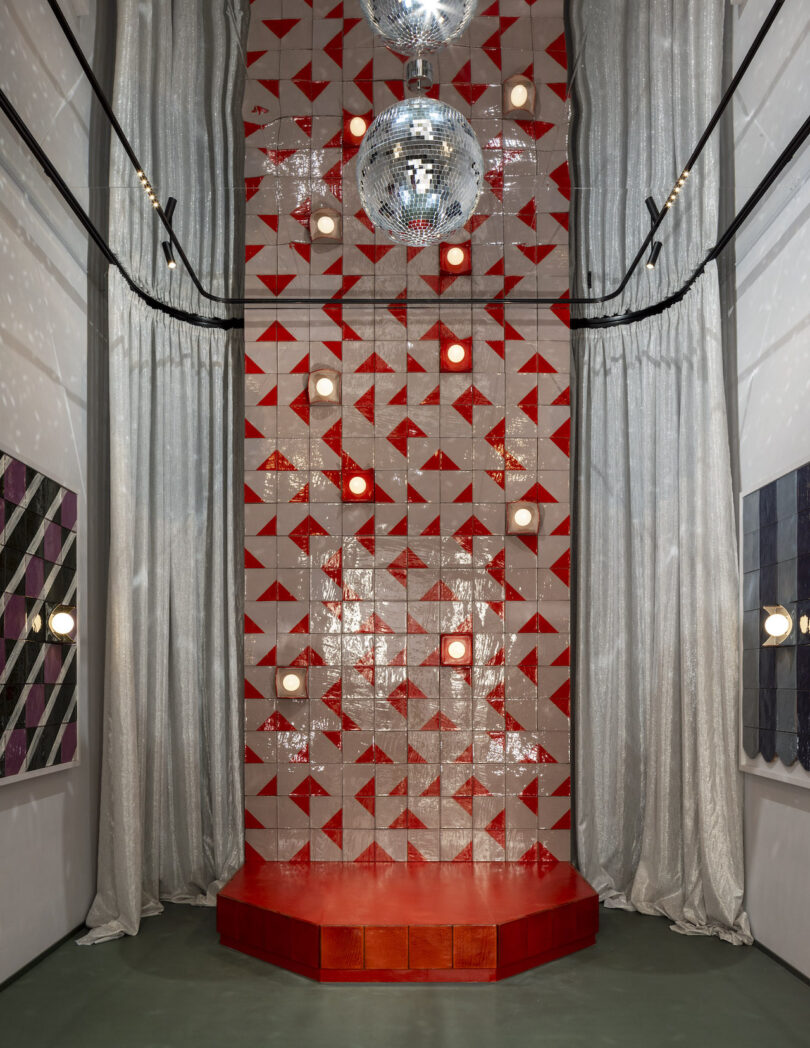 A small red stage is set against a red and white geometric tiled wall, with disco balls above and metallic silver curtains on either side