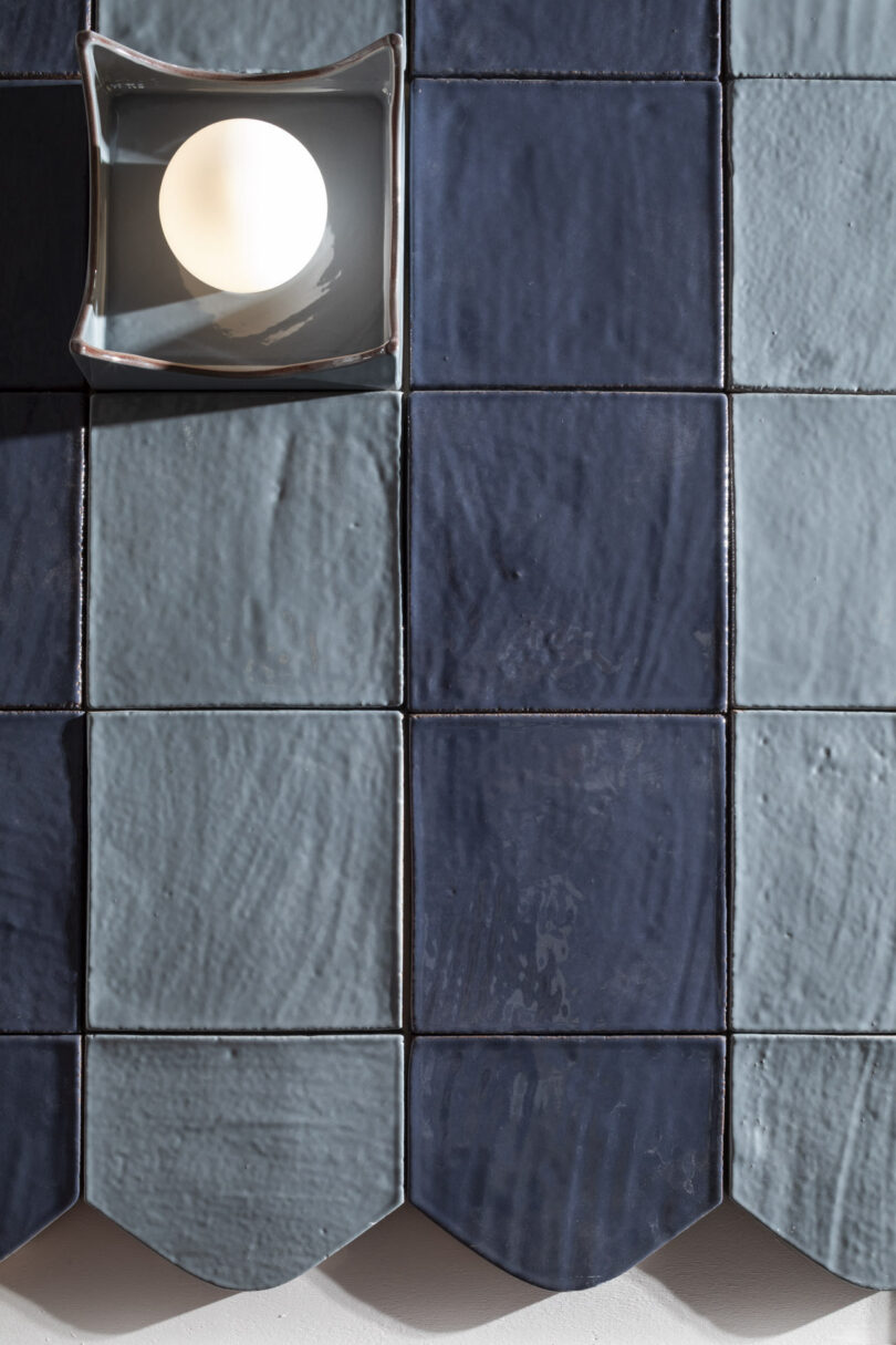 A lit round bulb in a square glass fixture sits on a grid of blue ceramic tiles with scalloped lower edges