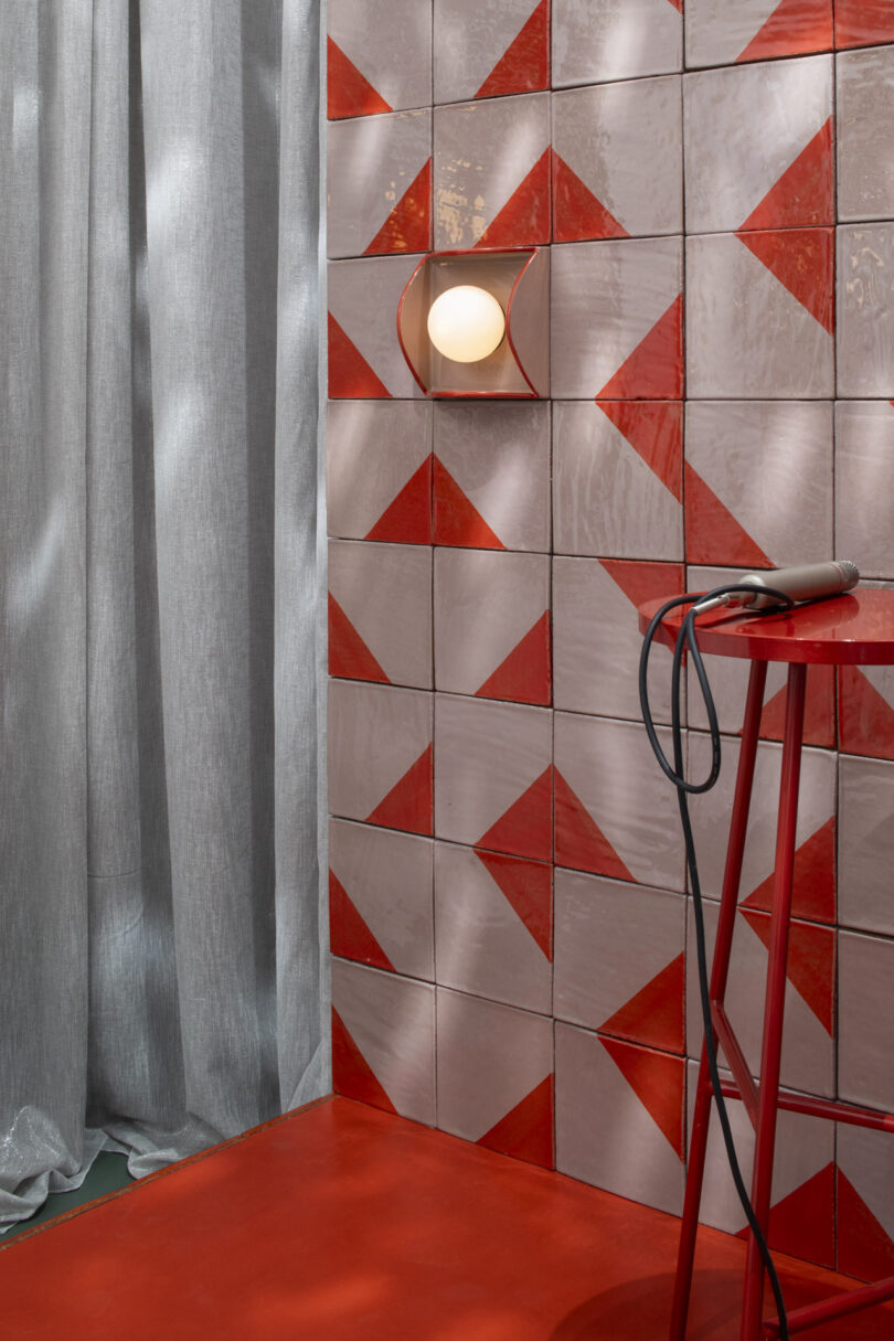 A red and pink geometric tiled wall with a round wall light, silver curtains, a red floor, and a red metal table holding a corded electronic device
