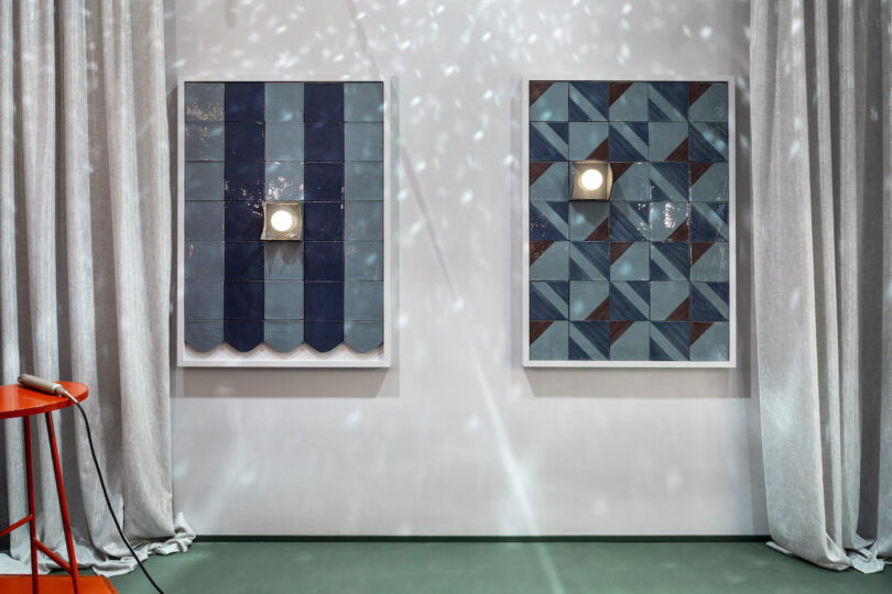 Two framed wall installations made of blue ceramic tiles with geometric patterns, each featuring a central light, are mounted between gray curtains on a white wall
