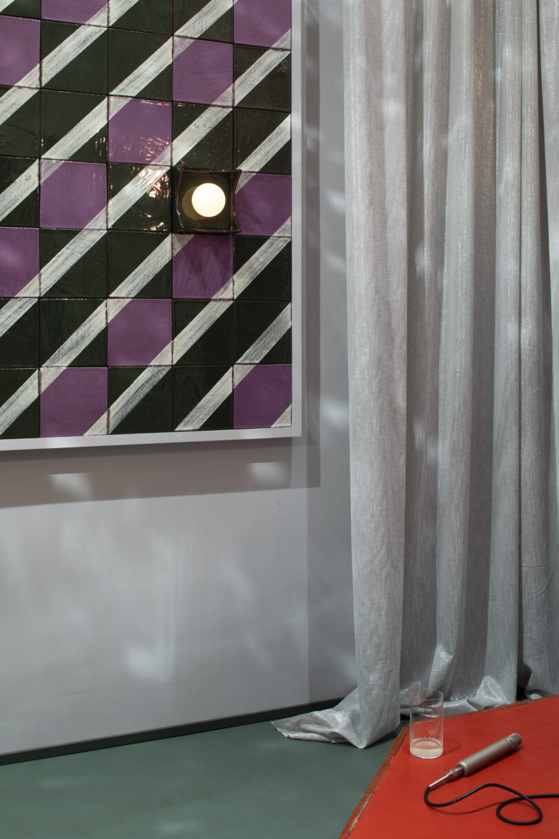 A tiled wall with a geometric green, purple, and white pattern, a mounted round light, a sheer curtain, and a red table with a glass of water and a microphone
