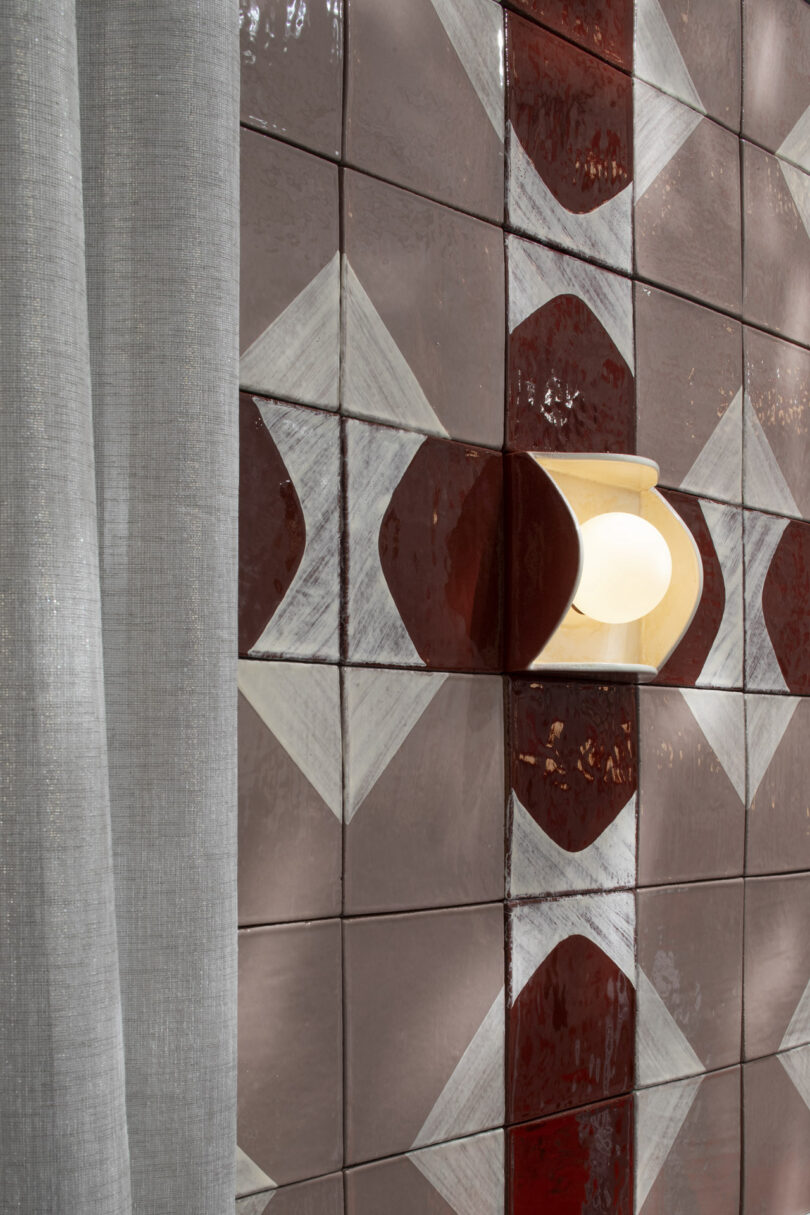 A wall with glossy, geometric-patterned red and white tiles next to a sheer gray curtain, featuring a mounted sconce light with a round bulb