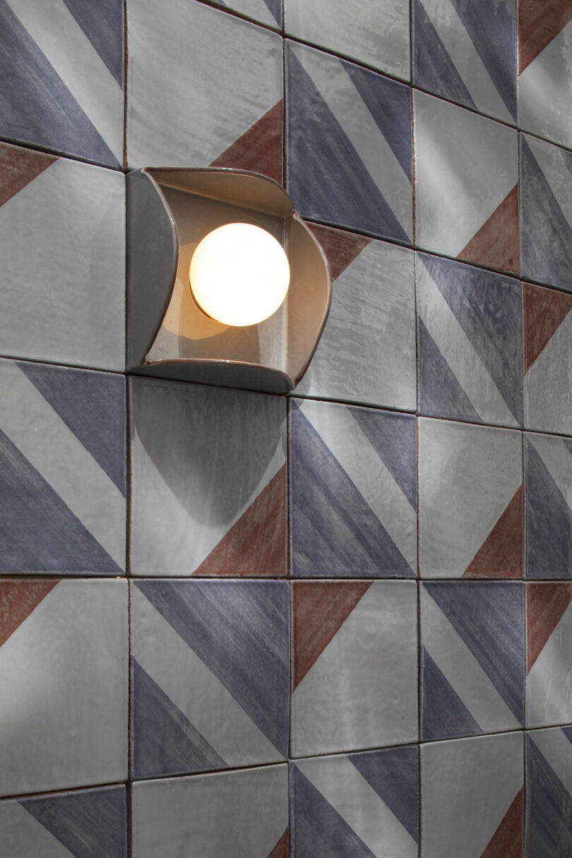 A round wall sconce is mounted on a tiled wall with a geometric pattern in shades of gray, blue, and red