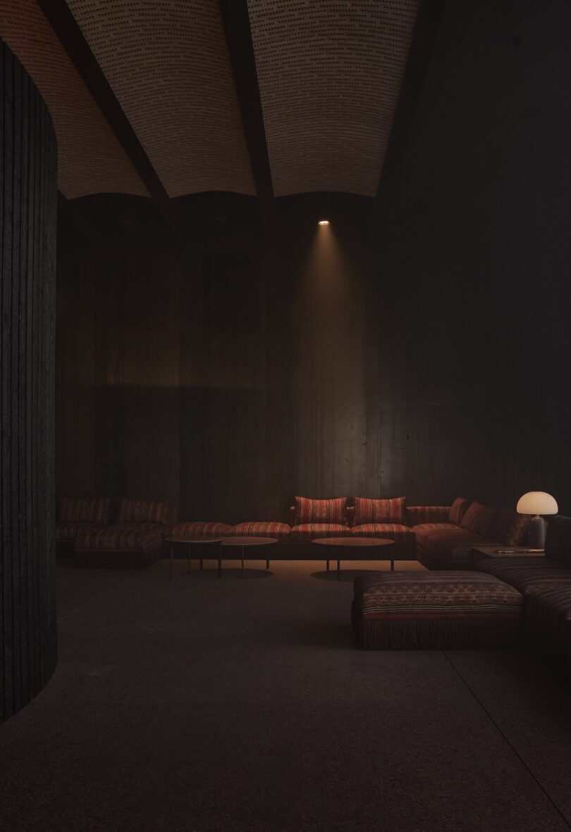 A dimly lit room with a curved sectional sofa, patterned upholstery, a round table lamp, and a single spotlight illuminating part of the seating area.