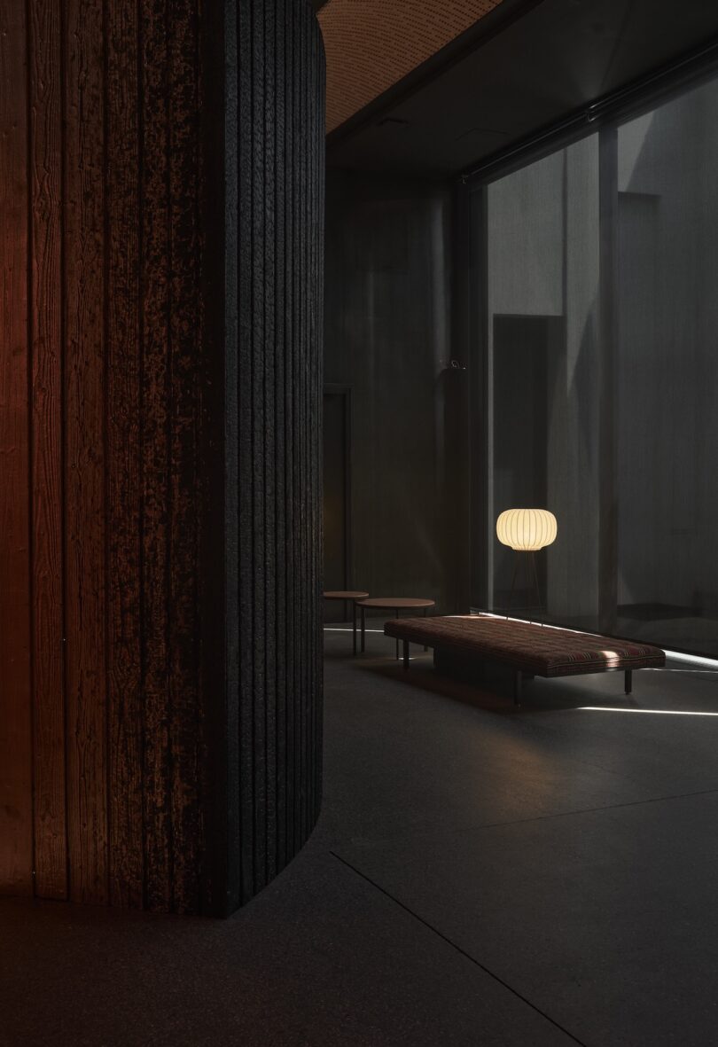 A modern minimalist interior with a textured dark wall, a low bench, and a round paper lantern lamp illuminated by soft natural light from large windows.