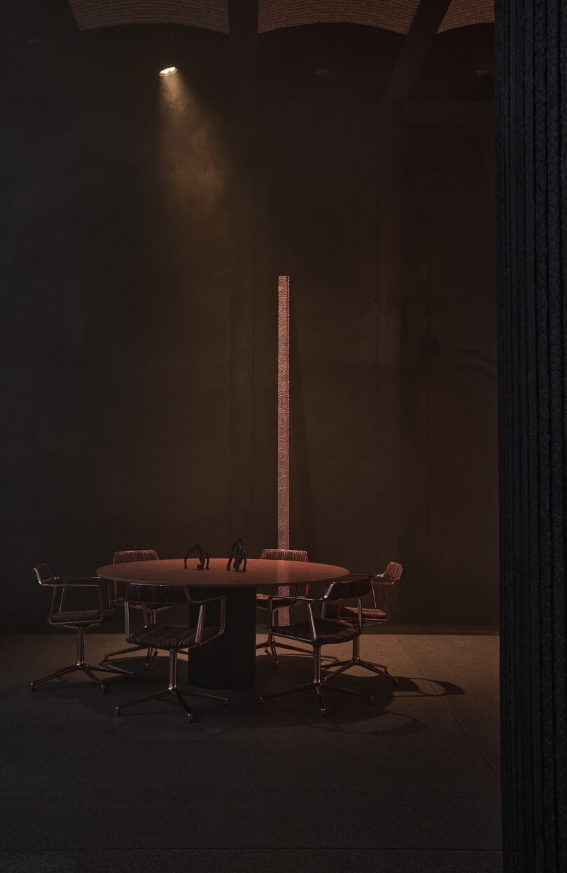 A round table with five chairs is lit by a spotlight in a dark, minimalist room; a tall, narrow pillar stands beside the table.