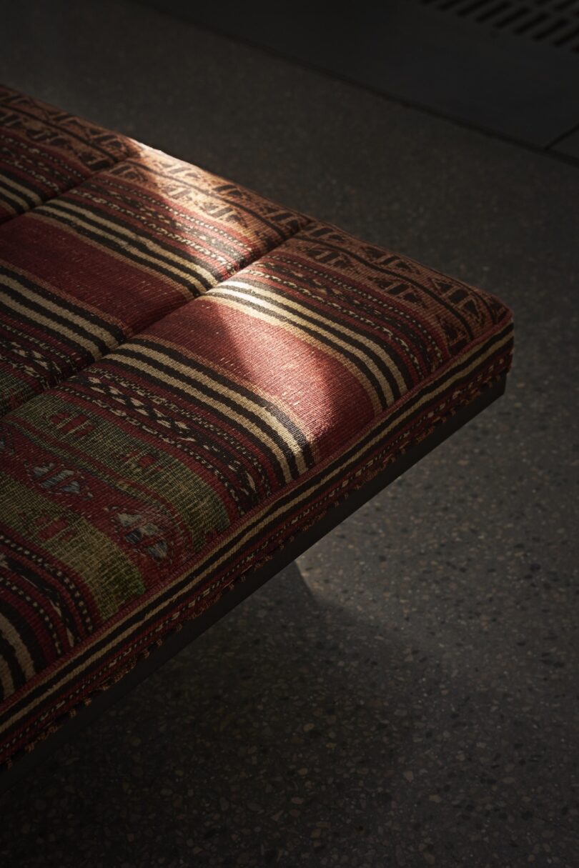 A patterned, upholstered bench seat with sunlight casting a diagonal shadow on it, set against a textured gray floor.