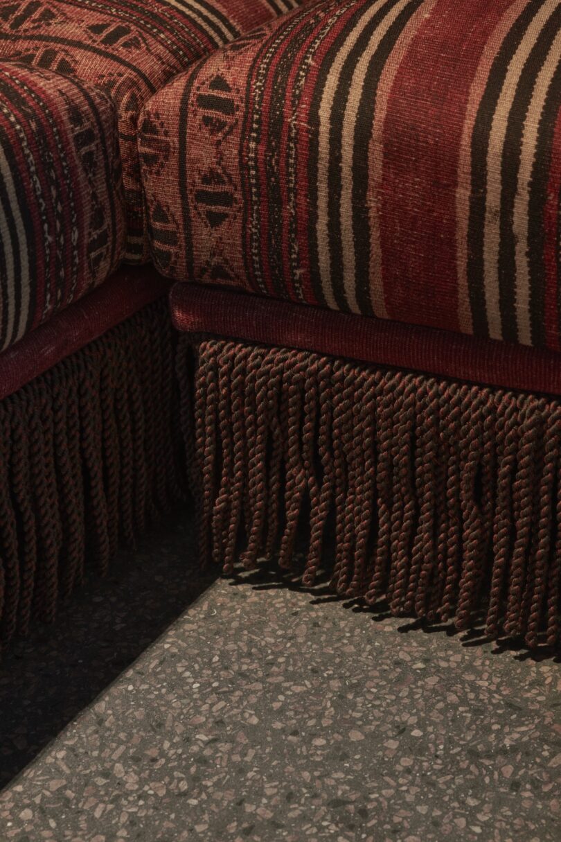 A close-up of two upholstered sofas with red patterned fabric, decorative fringe at the bottom, and a speckled terrazzo floor beneath.