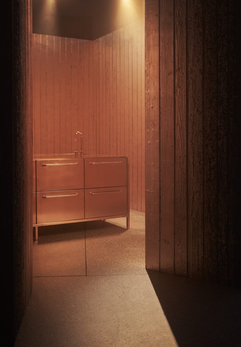 A minimalist bathroom features a metallic vanity with a faucet, set against wooden panel walls and stone flooring under warm, ambient lighting.