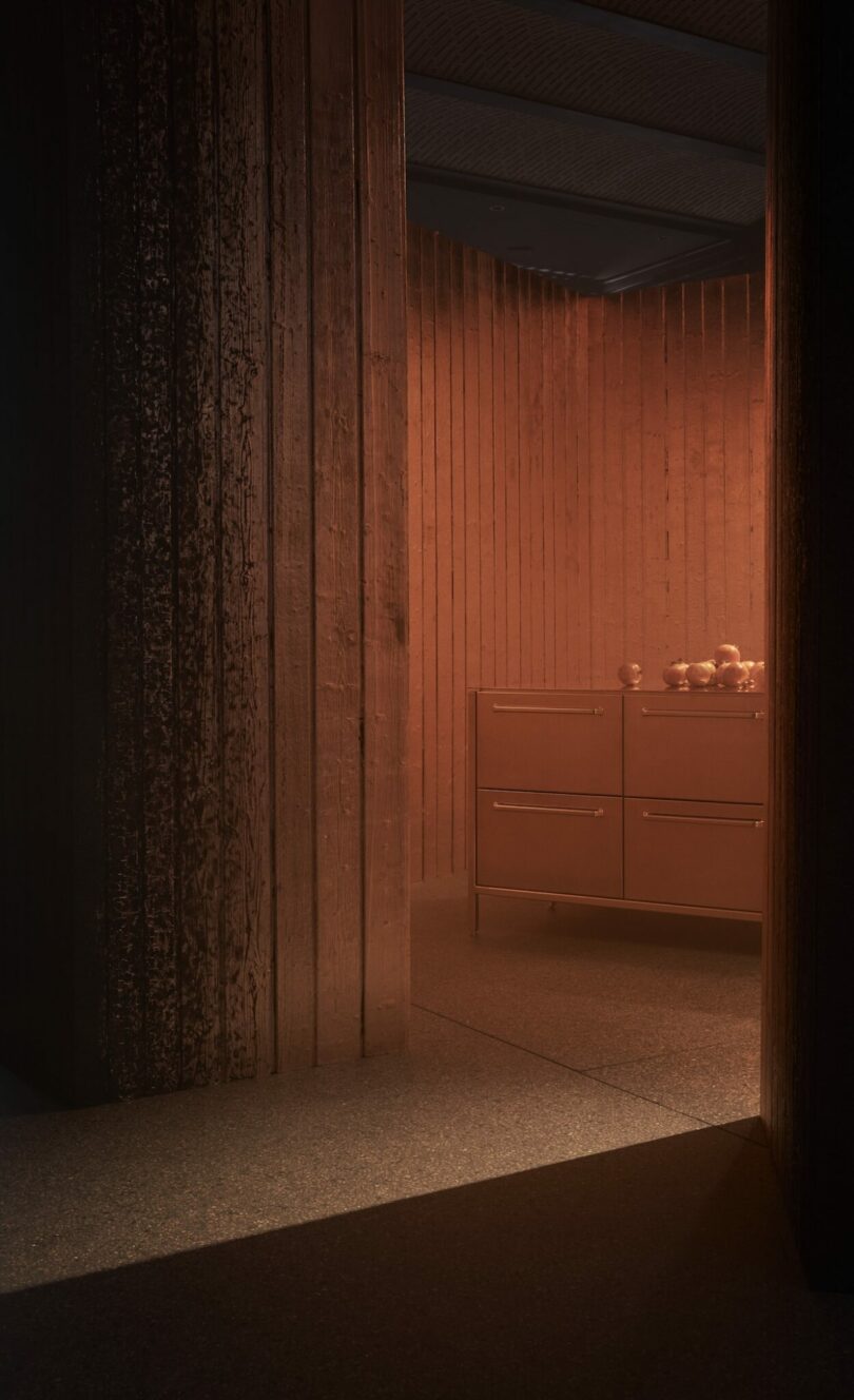A dimly lit room with textured wooden walls, a cabinet with drawers, and a bowl of fruit on top, illuminated by warm, orange lighting.