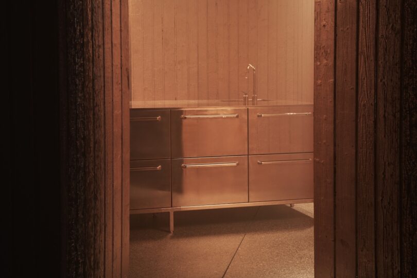 A stainless steel kitchen cabinet with drawers and a sink is seen through a doorway in a dimly lit room with wooden walls.