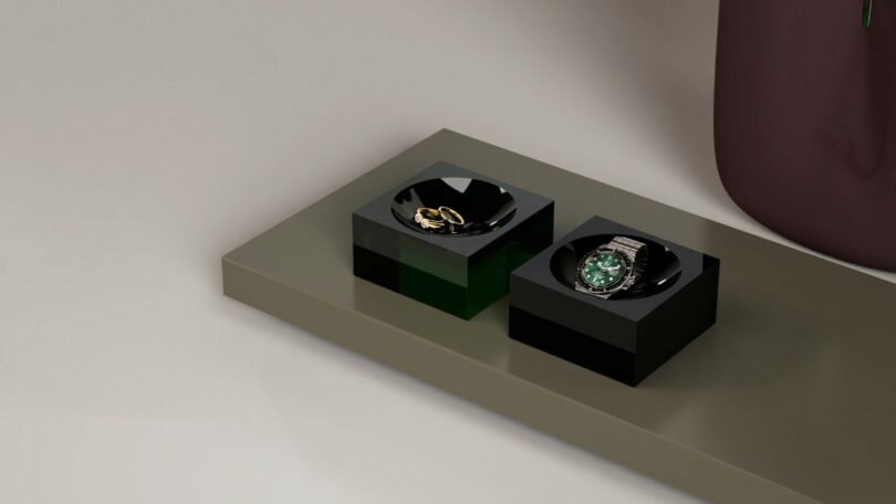 Two black square trays on a gray surface, one holding gold rings and the other holding a silver wristwatch with a green face