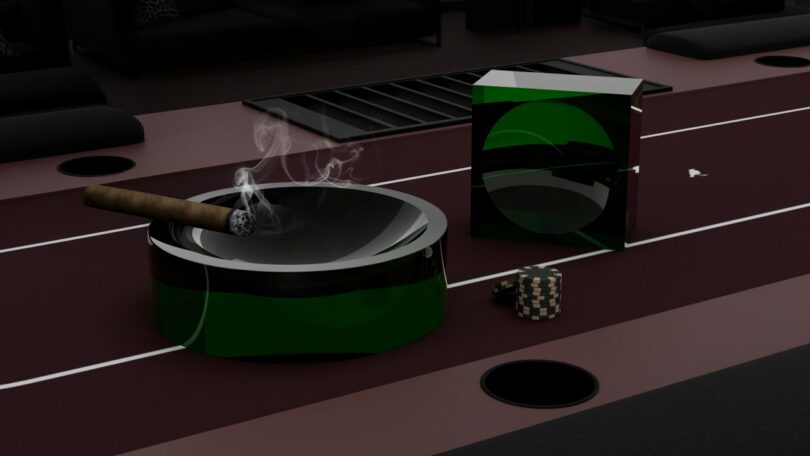 A lit cigar rests in a green and black ashtray beside a matching box and a small checkered object on a maroon table, with smoke rising from the cigar