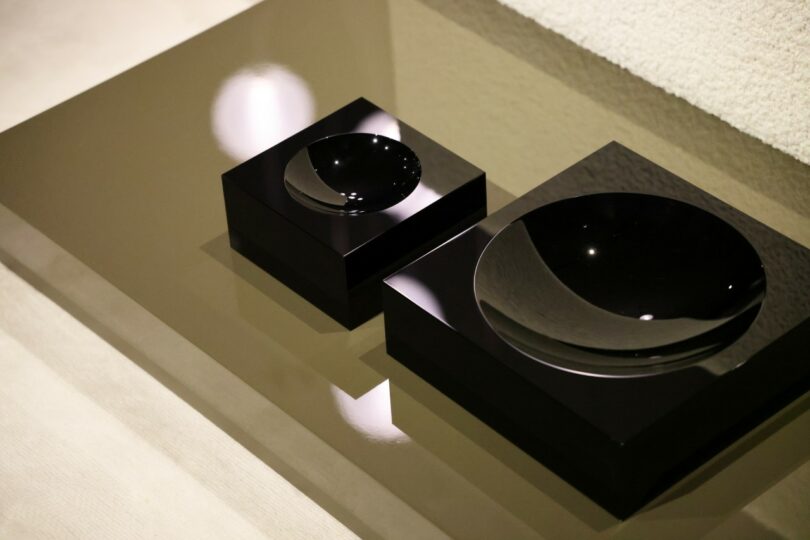 Two glossy black square objects with concave centers are placed on a reflective brown surface next to a textured white area