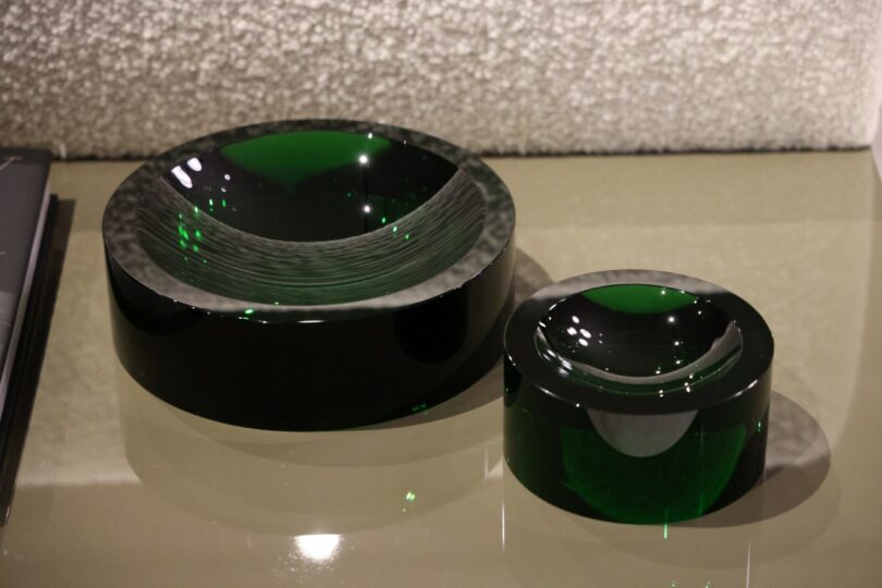 Two dark green glass bowls of different sizes are displayed on a reflective surface against a textured white wall
