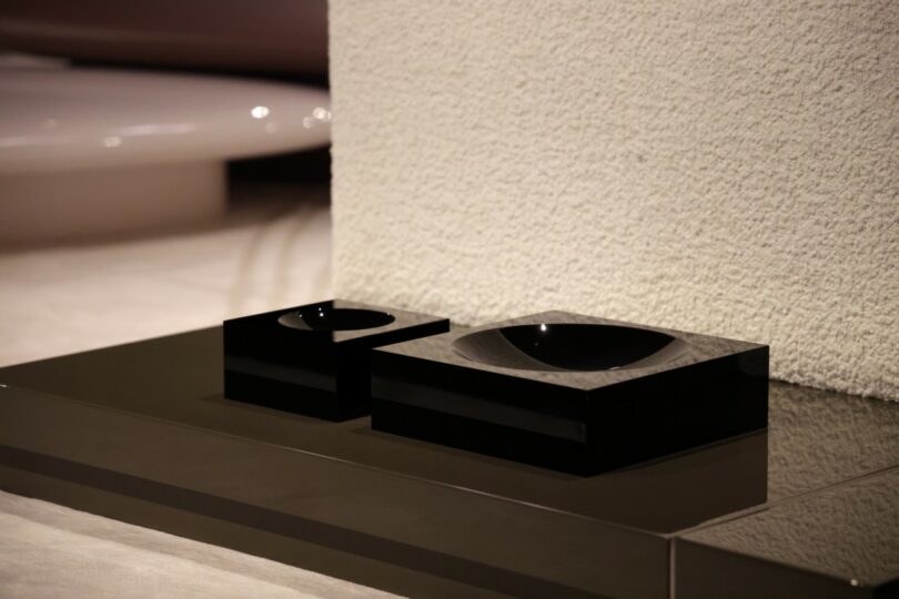 Two black, square, glossy bowls—one larger, one smaller—rest on a reflective dark surface next to a textured white wall