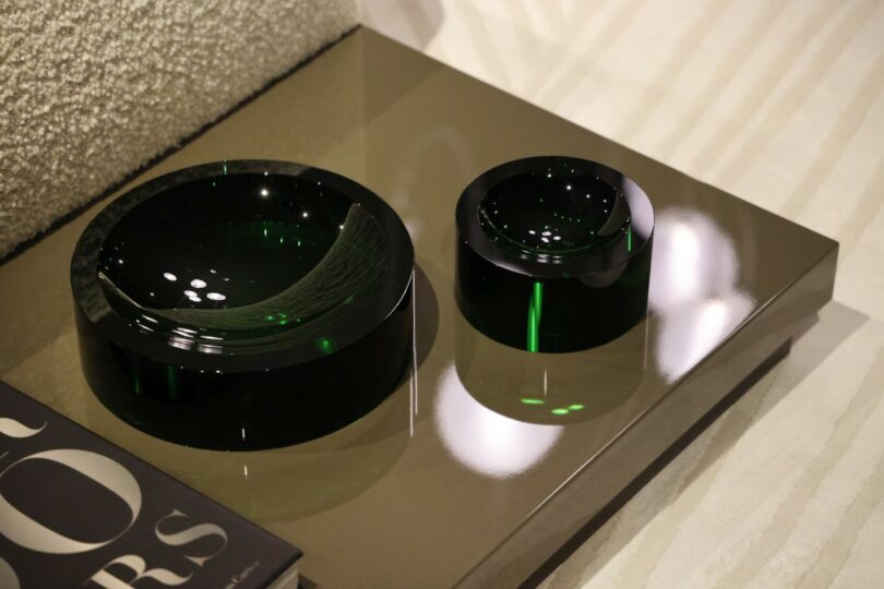 Two round, dark green glass bowls sit on a glossy beige table next to a partially visible magazine with large white text on the cover