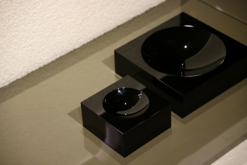 Two black, glossy, round dishes rest on matching square black bases, placed side by side on a reflective beige surface near a textured white wall