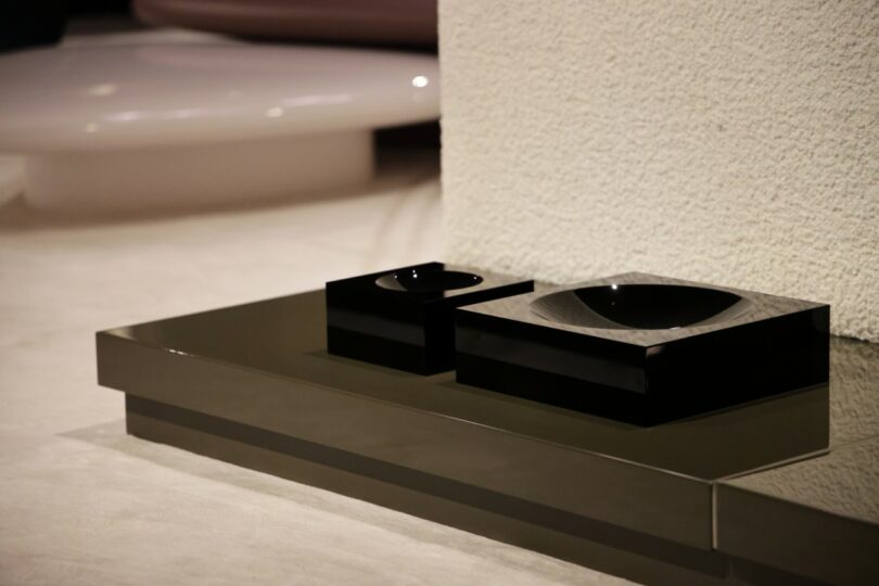 Two glossy black geometric bowls sit on a reflective surface near a textured white wall, with a large round object blurred in the background