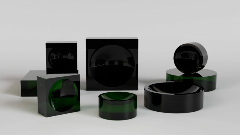 A group of glossy black and dark green geometric shapes, including cubes, cylinders, and spheres, displayed on a plain light gray background