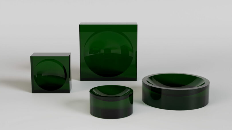 Four dark green, glossy geometric objects—two cubes and two cylinders—each with a hemisphere indented into their surfaces, displayed on a plain white background