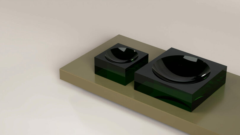 Two dark green and black square glass objects, one large and one small, sit on a beige rectangular platform against a plain light background