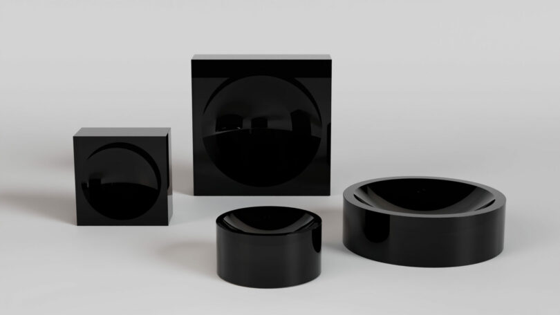 Four glossy black geometric objects, including two cubes and two cylinders, are arranged on a neutral surface with soft lighting